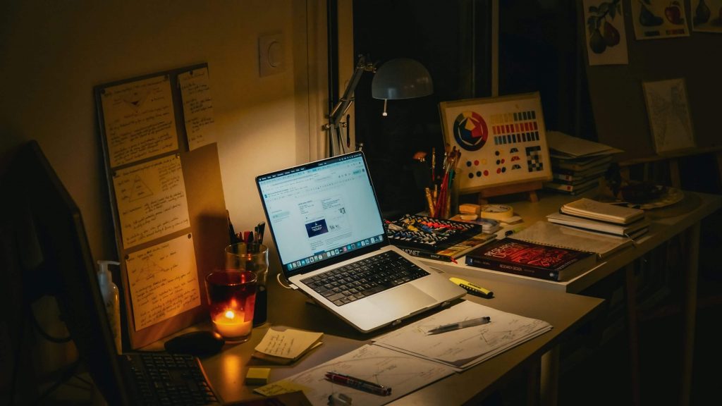 A desk workspace at night with a laptop, notebooks, sketches, art materials, and study notes under a desk lamp.