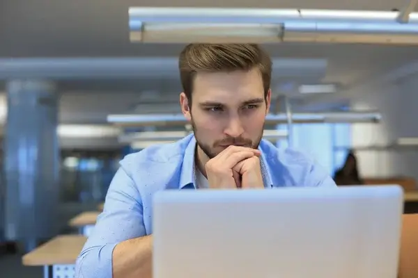 A man seated at a desk, concentrating on a laptop screen in a modern office or study environment.