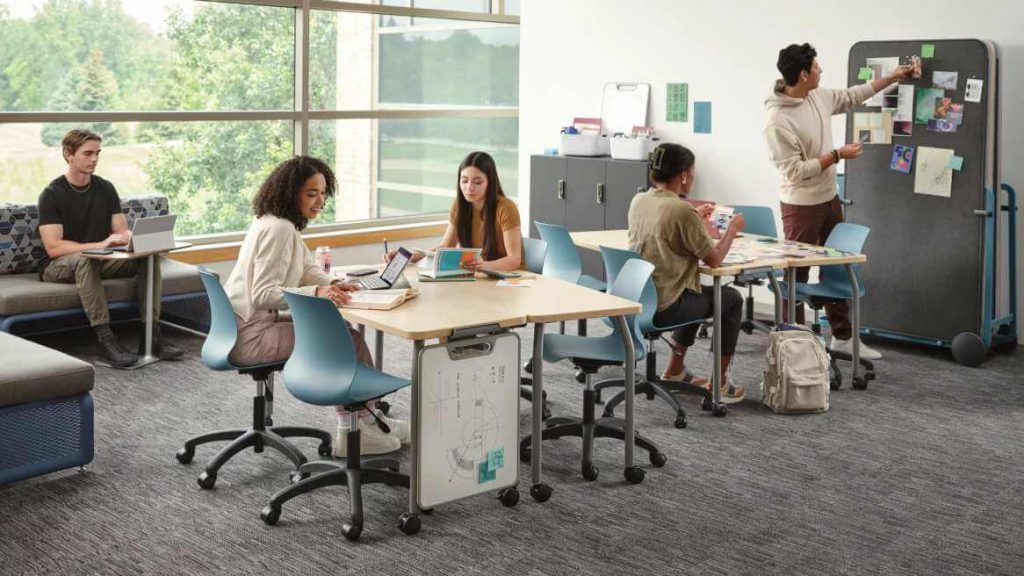A group of young people working together in a collaborative learning space with tables, laptops, notebooks, and a presentation board.