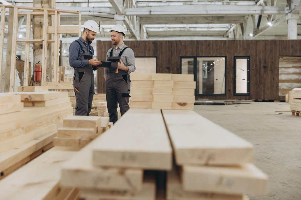 Two construction professionals reviewing plans in a workshop surrounded by wooden materials, representing teamwork and practical application of skills.