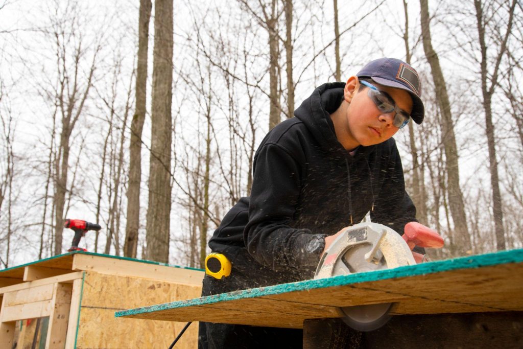 Young person using a circular saw to cut wood outdoors, demonstrating hands-on construction skills and practical learning.