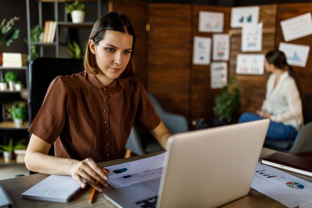 Woman working on a laptop reviewing charts and documents in an office, representing planning, strategy, and analytical thinking.