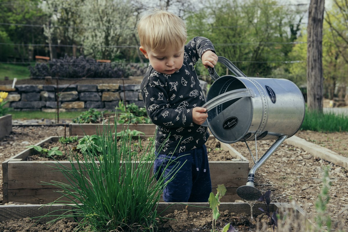 Child standing outdoors beside a watering can, ready to help with a garden task